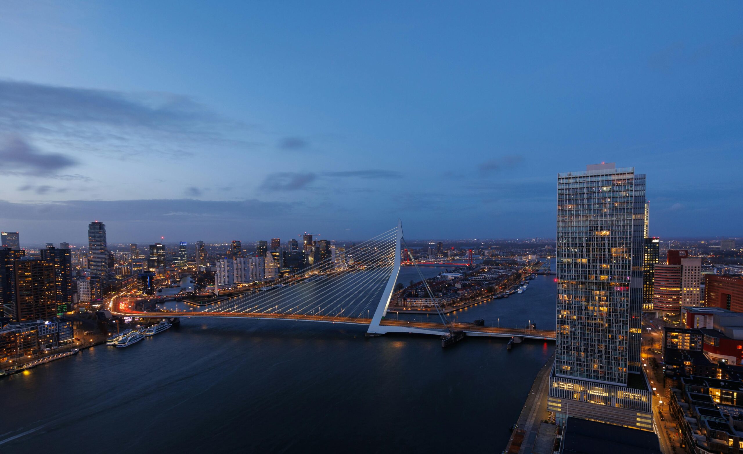 Stunning aerial view of the illuminated Erasmus Bridge and Rotterdam skyline at dusk.